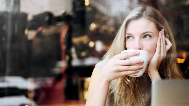 View through the window of charming young woman look out the window on snow city street enjoying cup of coffee or tea indoors Blond lady relaxing while sitting in cafe alone
