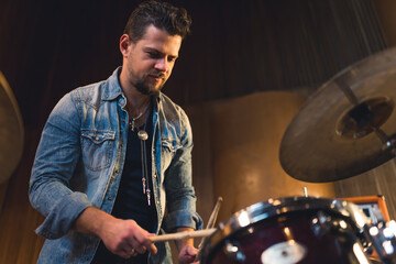 a young man with a beard wearing a jeans jacket holding sticks and playing drums in the studio....