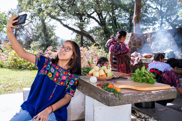 Naklejka na ściany i meble Chica haciendose una selfie con el telefono celular. Familia cocinando en una estufa de leña al aire libre. Familia latina, hispana. 