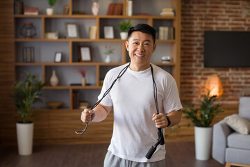 Excited asian middle aged man standing with skipping rope hang around his neck, ready to training at home in living room