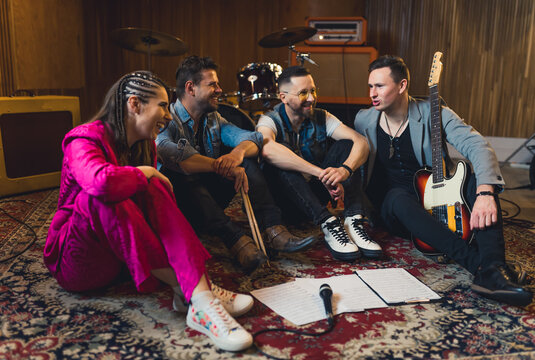 music band members sitting on the floor and talking cheerfully, drums and musical devices in the background, studio. High quality photo