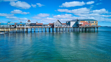 Aerial side profile of long old wood pier covered in shops in Maine with ocean and blue sky