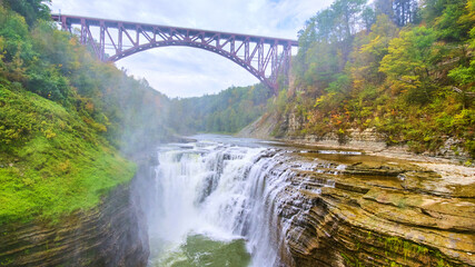 Waterfall cutting through cliffs from drone with steel maroon bridge overhead