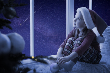 Cute little girl in Santa hat sitting on windowsill and looking at shooting star in beautiful night...