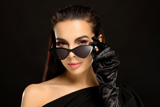Portrait of beautiful young woman in sunglasses and elegant evening gloves on black background
