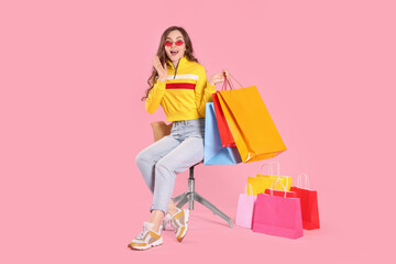 Obraz premium Emotional woman in stylish sunglasses holding many colorful shopping bags on armchair against pink background
