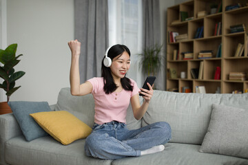 Excited chinese lady in wireless headphones looking at smartphone, making victory gesture, sitting on sofa