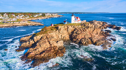Waves crash over rocks aerial over Maine island with lighthouse and view of homes on mainland