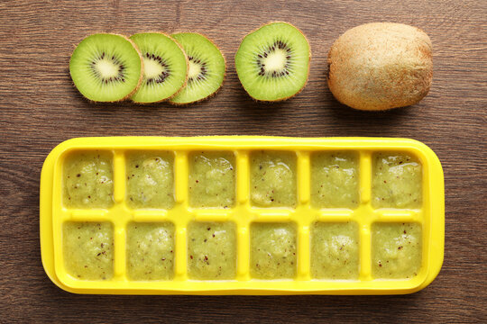 Kiwi puree in ice cube tray and fresh kiwi fruits on wooden table, flat lay. Ready for freezing