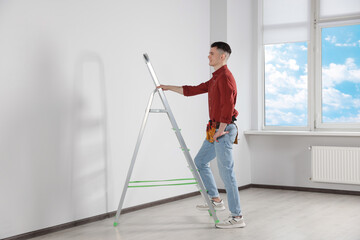 Young handyman with stepladder and tool belt working in room