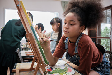 African American girl concentrates on acrylic color picture painting on canvas with students group...