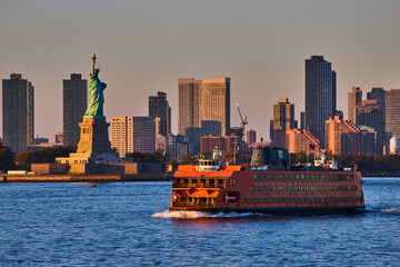 Large Staten Island Ferry passing by Statue of Liberty in golden hour of New York City with skyline...