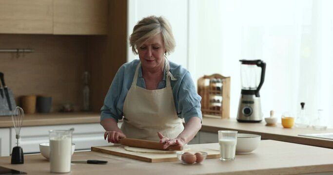 Elderly 55s woman prepares pastries in kitchen, attractive smiling mature female wear apron hold rolling-pin flattening prepared dough makes homemade pie. Family recipe, natural ingredient for cookery