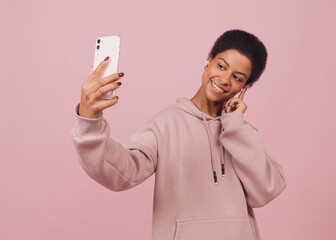 Beautiful smiling black girl shooting selfie on front camera. Close-up portrait of happy cheerful african american woman having video-call and holding smart phone in hand over pink background