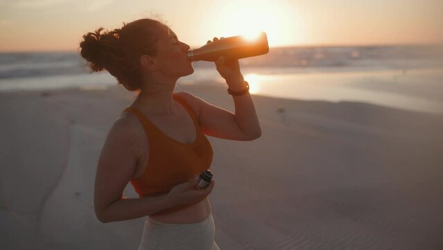 fit sports woman in fitness clothes at the beach at sundown drinking water.