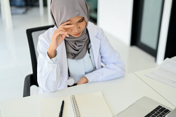 Stressed Asian Muslim female doctor concentrating on her medical research, working at her desk