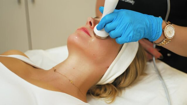 Indoor closeup trait of attractive blonde adult caucasian woman in white tank top during microdermabrasion procedure performed by unrecognizable beautician in black uniform. High quality 4k footage