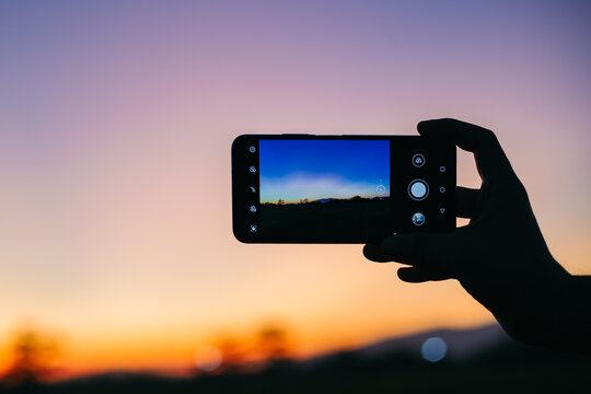 Close-up of hand with a phone taking photos of a bright sunset and mountain in countryside landscape view