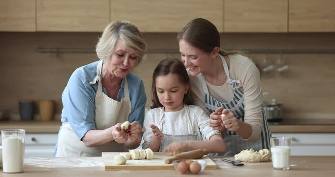 Little cute girl her older granny and loving young mother in aprons cooking together, forming prepared dough making homemade delicious buns, enjoy communication, time together and cookery in kitchen