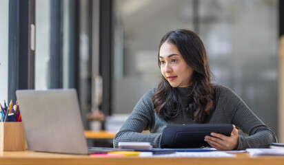 Freelancer Happy business Asian woman in knitwear taking notes at laptop sitting at desk office, finance concept.