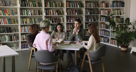In university library group of international students discuss task seated at table with heap of textbooks, share ideas, prepare for high school exams or admission, make exercises, engaged in teamwork