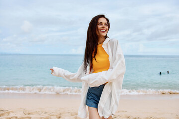 Fototapeta na wymiar Portrait of a happy woman smile with teeth with long hair brunette walks along the beach in a yellow tank top denim shorts and a white shirt by the sea summer travel and feeling of freedom, balance