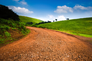 Fototapeta na wymiar Rugged muddy road passing through beatiful green grass rolling hills with fluffy clouds.
