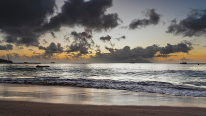 Obraz premium Twilight after sunset on a tropical beach. There are picturesque clouds in the orange-lit sky. Outlines of the island and yachts on the horizon. Foam of waves and reflection on wet sand. Seychelles. 