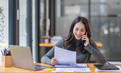 Cheerful business Asian woman freelancer making telephone call share good news about project working in office workplace, business finance concept.