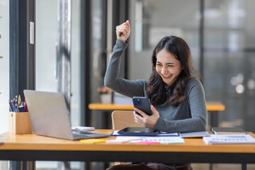 Happy excited successful business asian woman triumphing in office, Portrait of a cheerful Asian businesswoman sitting at the table in office, 	