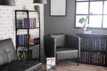 Shelf unit with books, sofa and armchair near grey brick wall in library interior