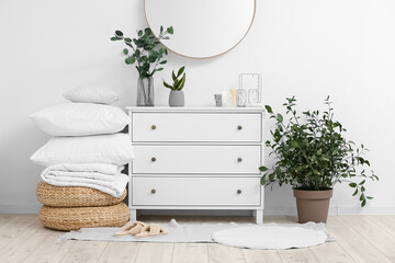 Chest of drawers with houseplants, bed sheets and poufs near light wall