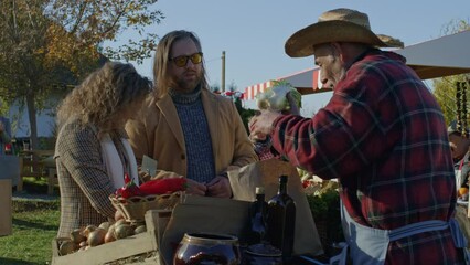 Family shopping on local farmers market. Holder of sales point offers own product and gives eco bag with fruits or vegetables. Wife pays for purchase by cash.