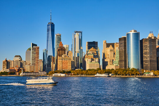 Ferry passing across southern Manhattan skyline from water with golden light hitting skyscrapers
