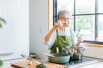家のキッチンで料理を作って味見するアジア人の高齢者女性（笑顔）
