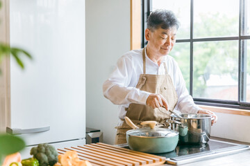 家のキッチンでフライパンを使って手料理を作るアジア人の高齢者男性（笑顔）
 - obrazy, fototapety, plakaty