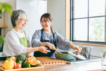 家のキッチンで一緒に料理・家事・自炊をする笑顔のアジア人高齢者女性と若い女性（家政婦・家事代行）

