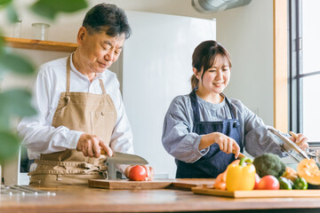 家のキッチンで一緒に料理・家事・自炊をする笑顔のアジア人高齢者男性と若い女性（家政婦・家事代行）

