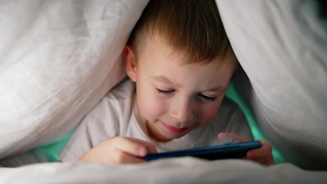 Baby boy lies under covers on bed and plays games on mobile phone. Child at leisure using modern wireless technologies and has rest. Watch videos on social networks. Problem of addiction to gadgets.