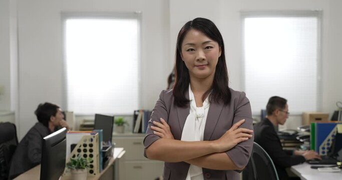 half length portrait of professional asian woman manager smiling at camera with folded arms in bright modern office with colleagues working at background.