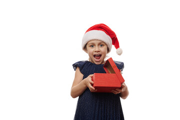 Cheerful child, lovely baby girl wearing Santa Claus hat, expressing positive emotions, happiness, joy and surprise, looking at camera while unpacking a happy Christmas present, on white background