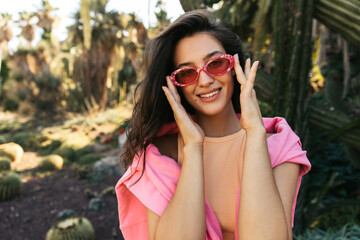 Beautiful young caucasian girl corrects her fashionable sunglasses, looking at camera with smile. Brunette wears top and sweater. Positive emotions concept.