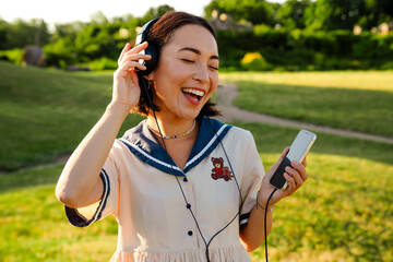 Asian girl singing while listening music with headphones and mobile phone in park