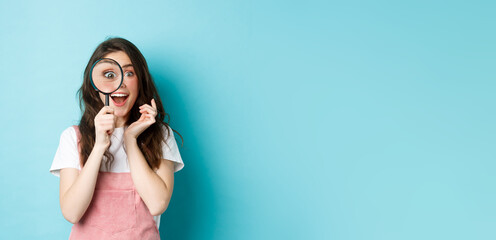 Happy young woman looking through magnifying glass with excited face, found or search something, standing over blue background - obrazy, fototapety, plakaty