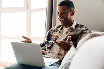 Positive african man having video call via laptop while sitting on couch at home