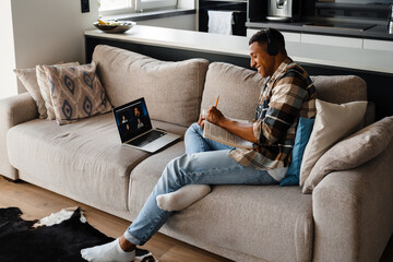 Obraz premium Afro man making online conference call on laptop and making notes while sitting on sofa