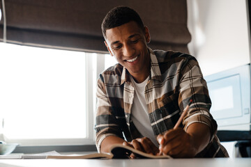 Cheerful african man writing down notes while sitting at table at home
