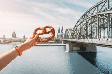 Traditional german snack - pretzel or bagel on the background of an Cologne cathedral building and bridge over Rhine river. Cuisine and tourism in Germany and Rhineland - obrazy, fototapety, plakaty