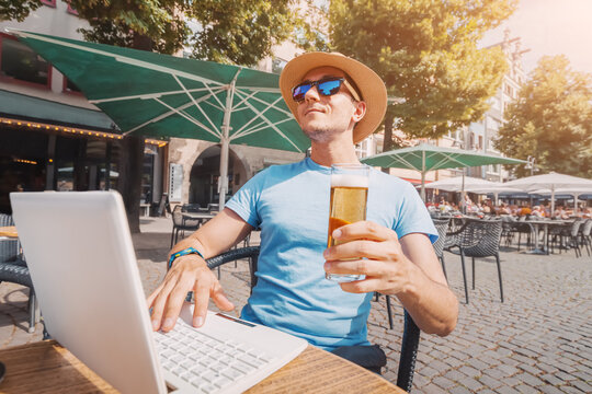 Happy man relaxes and drinks a glass of beer in a pub in the fresh air and works or communicates via laptop
