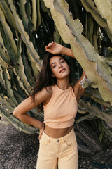 Top view of calm young caucasian girl leaned on big cactus, stands in botanic park alone. Brunette wears casual spring clothes. Concept of relaxing.